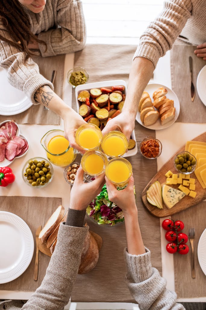 Top view of friends toasting with orange juice during breakfast with a variety of foods.